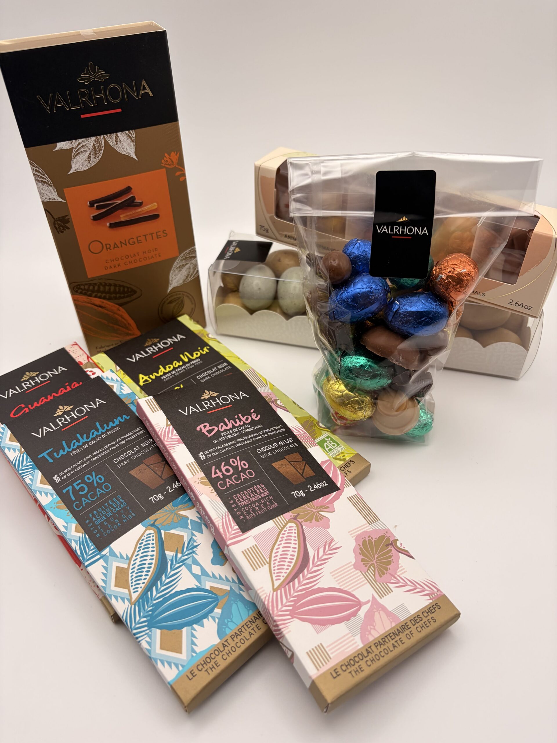 Assortiment de produits Valrhôna : tablettes de chocolat Tulakalum 75%, Bahibé 46%, coffret d'orangettes et œufs en chocolat colorés sur fond blanc.