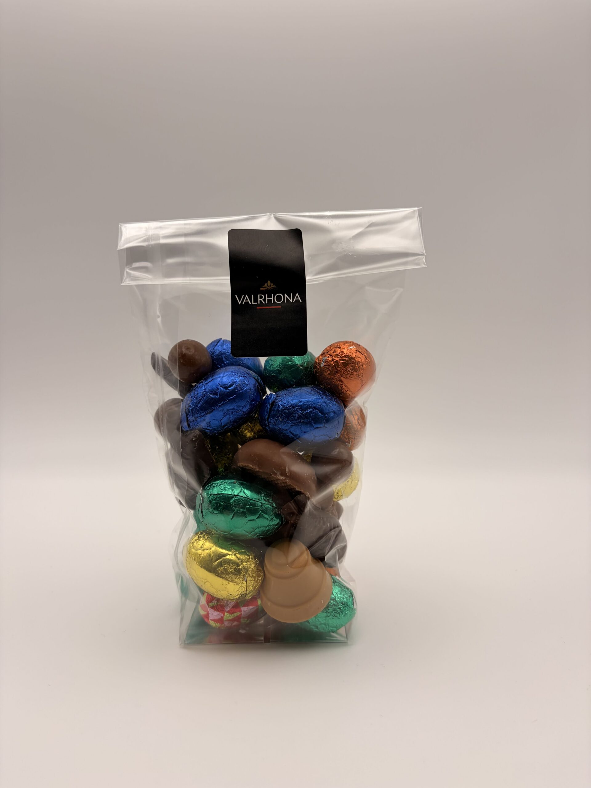 Un sachet transparent fermé par une étiquette noire Valrhôna, rempli d'œufs en chocolat emballés dans des papillotes colorées bleues, vertes, oranges et dorées.