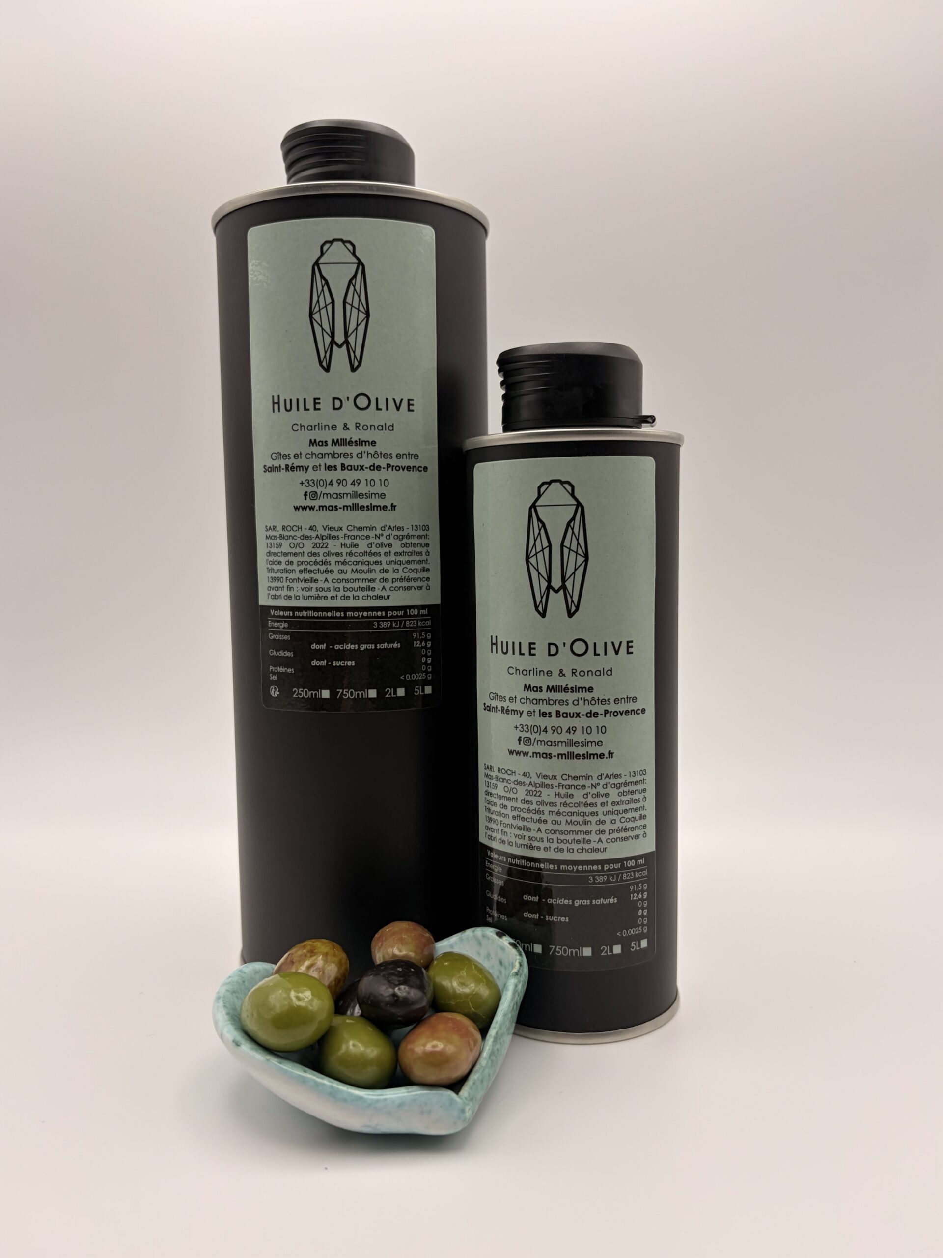 Deux bidons métalliques noirs d'huile d'olive vierge extra du Mas Millésime (formats 250ml et 750ml) ornés d'une cigale stylisée, posés derrière un petit ramequin en forme de cœur rempli d'olives vertes et noires.