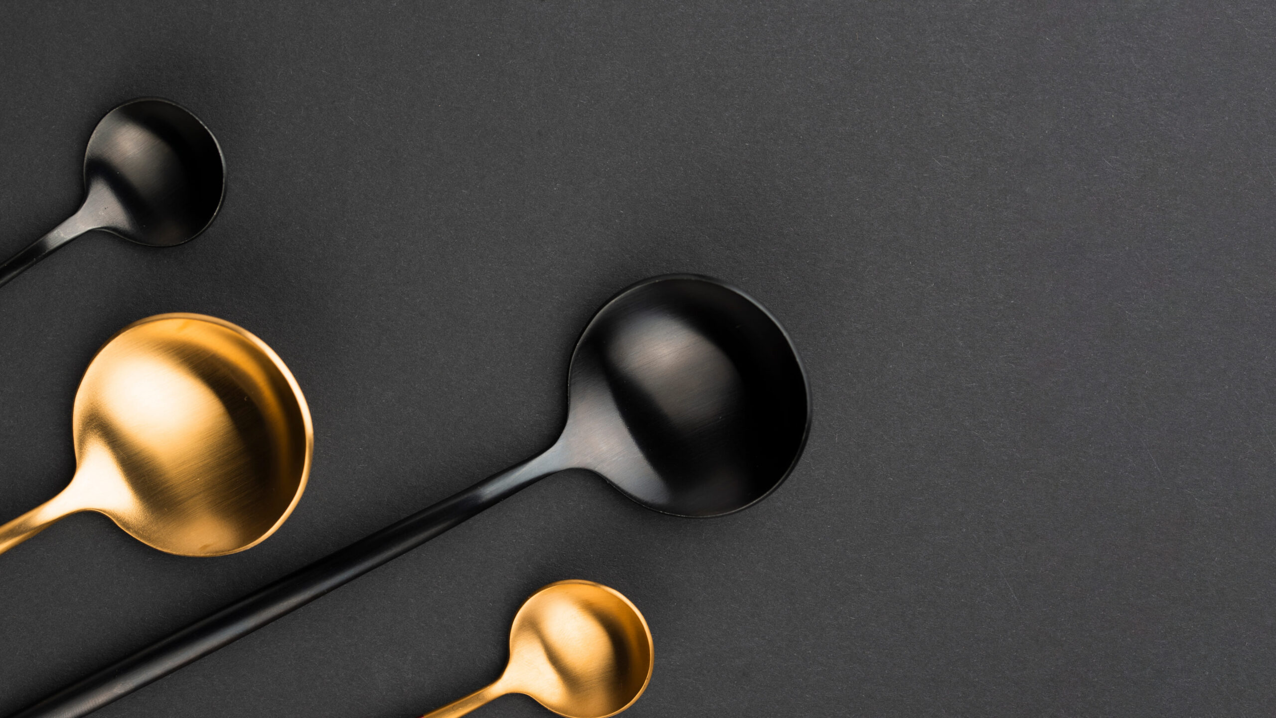 Quatre cuillères design de couleur noir mat et doré, disposées de manière asymétrique sur un fond texturé gris anthracite foncé.