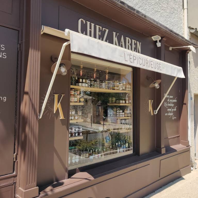 Façade en bois marron de la boutique d'épicerie fine Chez Karen - L'Épicurieuse à Saint-Rémy-de-Provence, arborant des lettres dorées, un store banne blanc déplié et une grande vitrine lumineuse laissant apercevoir des étagères remplies de bocaux artisanaux.