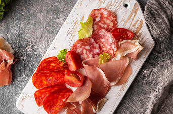 Assortiment de charcuterie artisanale comprenant du jambon cru, du saucisson et du chorizo, présenté sur une planche en bois blanc avec des tomates cerises.