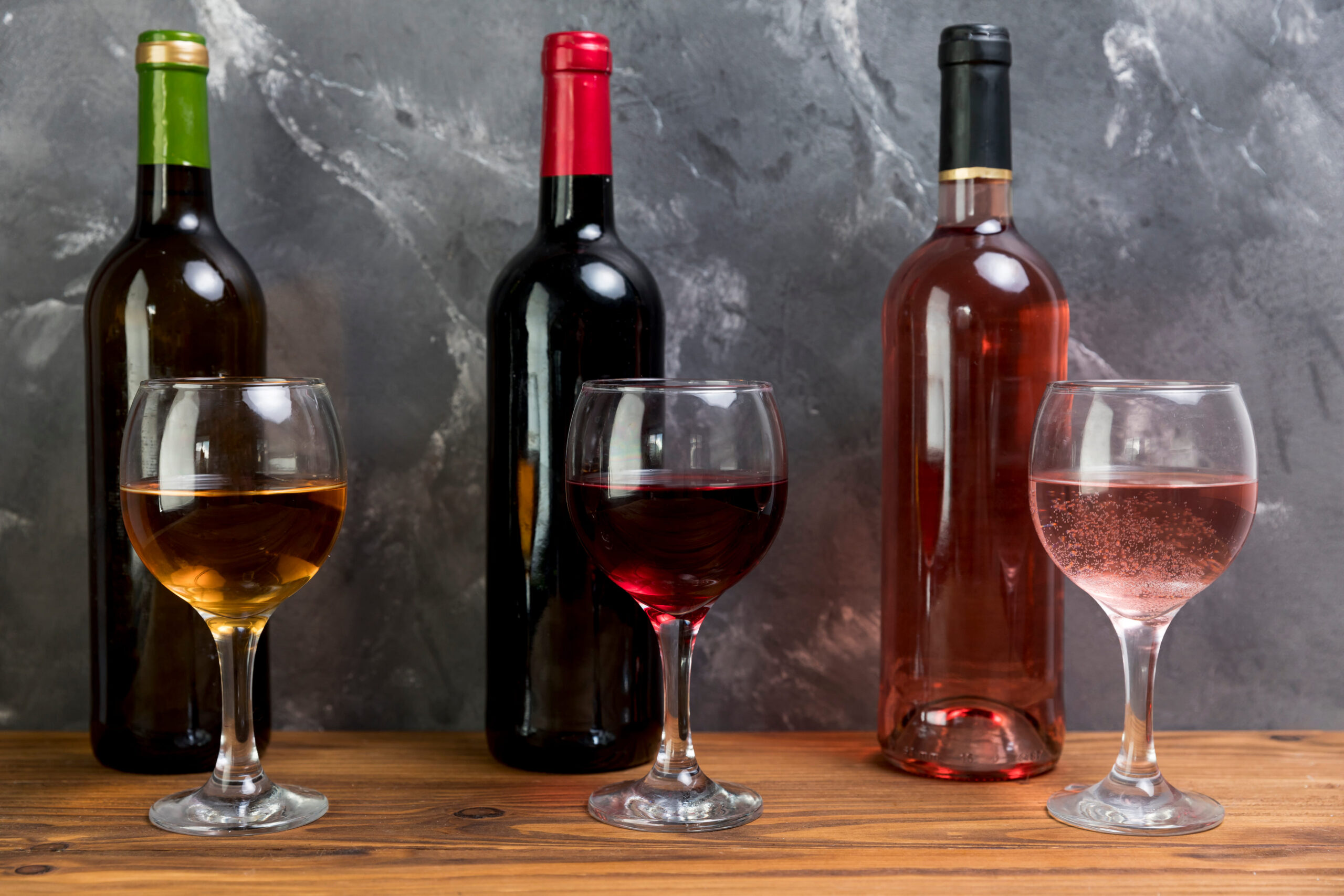 Sélection de Vins et Spiritueux d’Exception – Chez Karen Alignement de trois bouteilles de vin (blanc, rouge et rosé) accompagnées de leurs verres remplis respectifs, posés sur un plateau en bois devant un mur gris texturé.